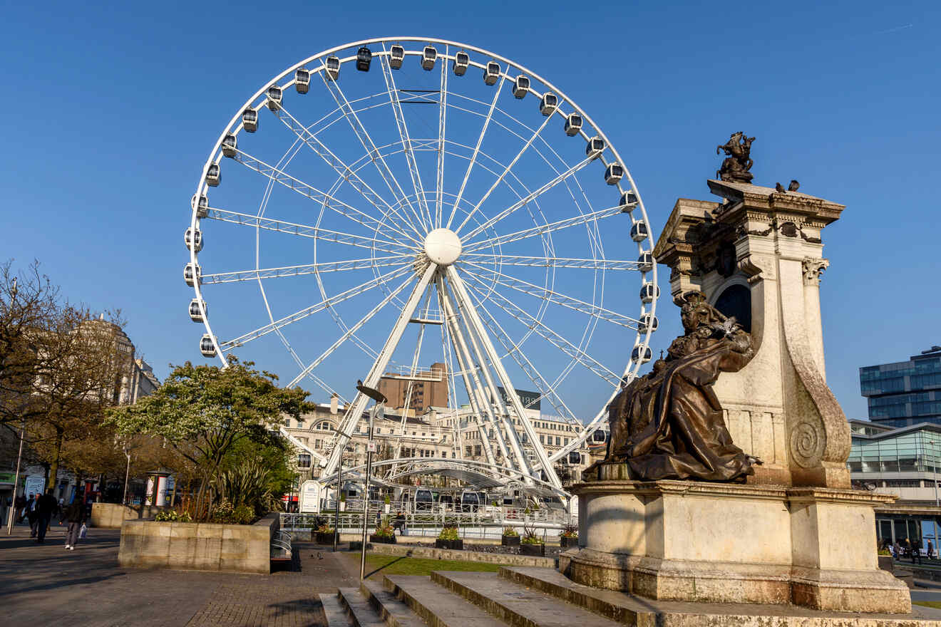 The Manchester Wheel, a large observation Ferris wheel located in the city center, with blue skies and the statue of Queen Victoria in the foreground